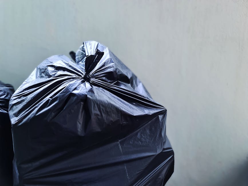 Close-up image of multiple black plastic rubbish bags filled and tightly tied, stacked together against a plain, light-colored wall. The bags are made of glossy plastic, reflecting light, with some sh